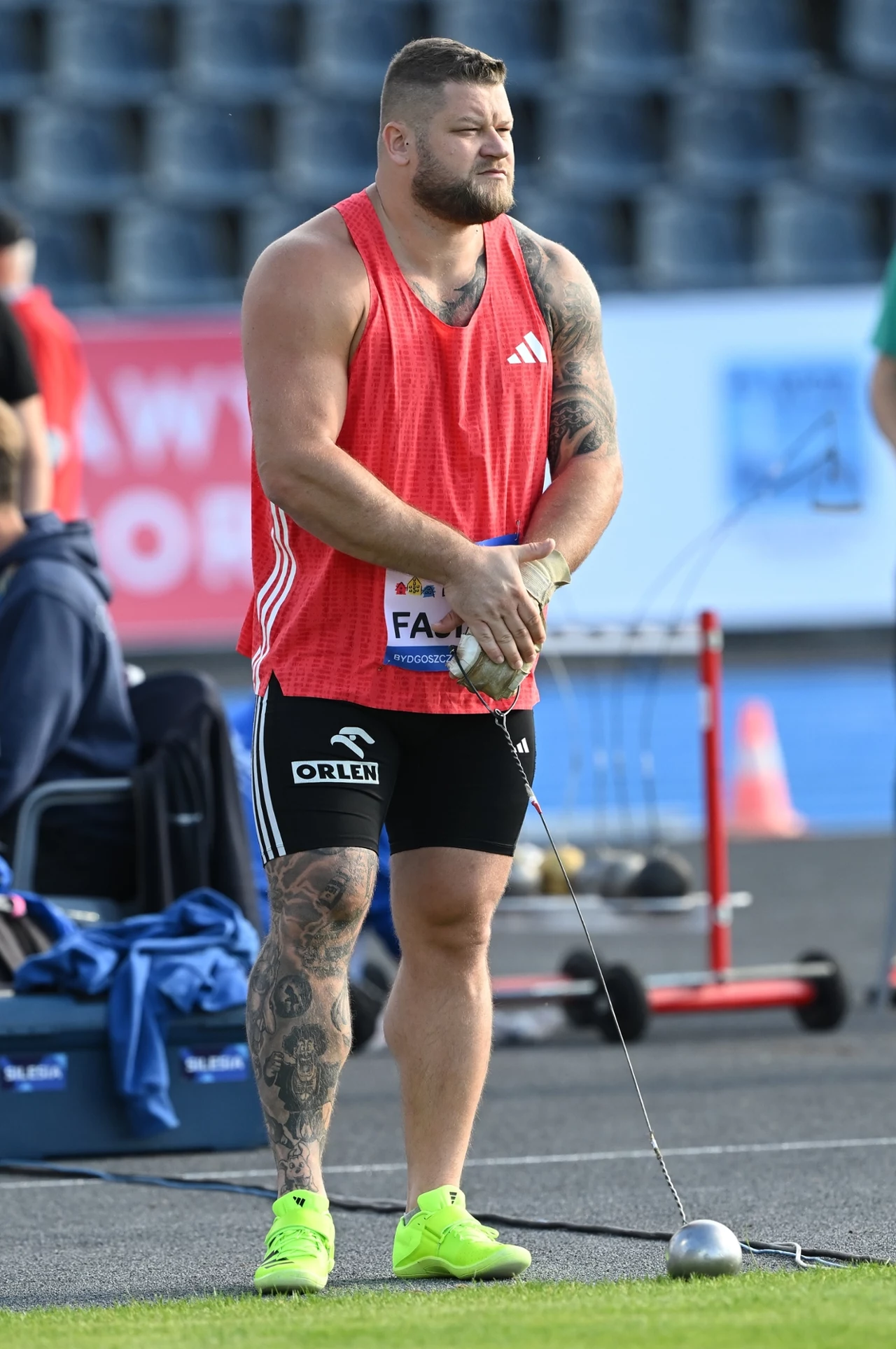 Atleta w charakterystycznej czerwonej koszulce i czarnych spodenkach przygotowuje się do rzutu młotem na stadionie lekkoatletycznym, trzymając młot w dłoniach, na tle pustych trybun.
