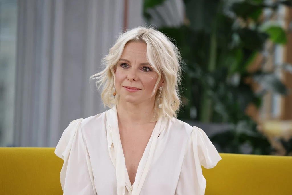 Alicja Janosz w 2023 r. Kobieta o blond włosach w eleganckiej białej bluzce siedzi na żółtej sofie, w tle rozmyte rośliny oraz wnętrze salonu.