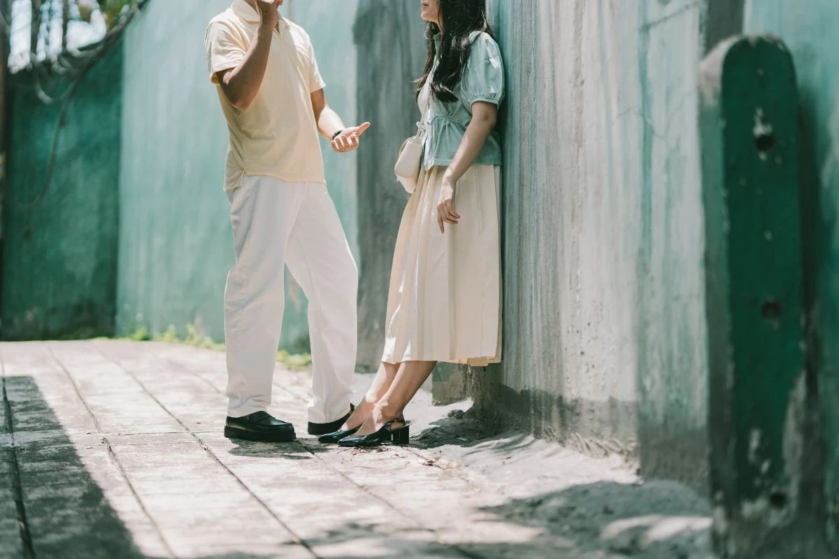 Nie tak łatwo znaleźć drugą połówkę Dwie osoby rozmawiające ze sobą, stojące obok wysokiego, szarego muru w zacienionym miejscu, jedna osoba wspiera się plecami o ścianę, druga gestykuluje rękami.