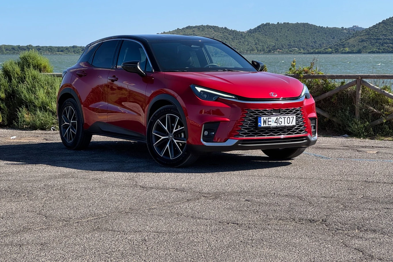 Czerwony nowoczesny samochód typu SUV zaparkowany na utwardzonym terenie, w tle widoczne jezioro, zielone wzgórza i drewniane ogrodzenie.