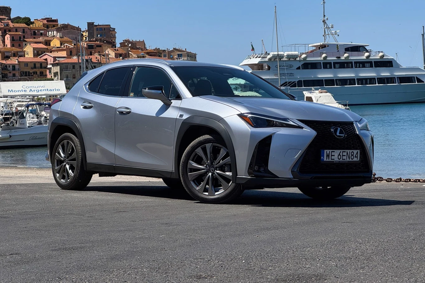 Srebrny samochód Lexus UX zaparkowany na tle portu z żaglówkami oraz luksusowym jachtem, w oddali widać kolorowe zabudowania nadmorskiego miasteczka.