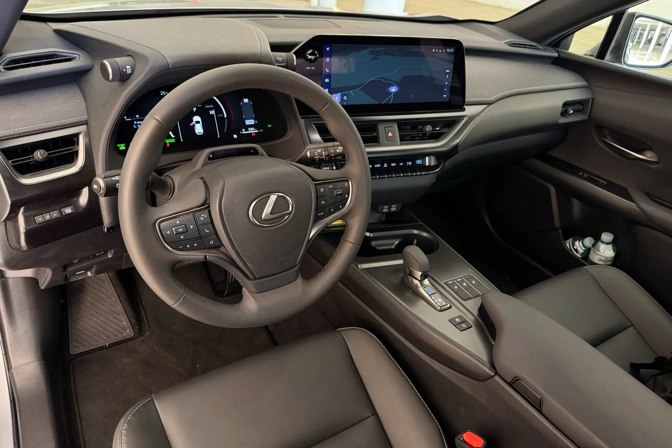 Wnętrze nowoczesnego samochodu Lexus z centralnie umieszczonym wyświetlaczem multimedialnym, cyfrowym panelem wskaźników oraz wielofunkcyjną kierownicą. Na fotelu pasażera widoczne dwie butelki wody.