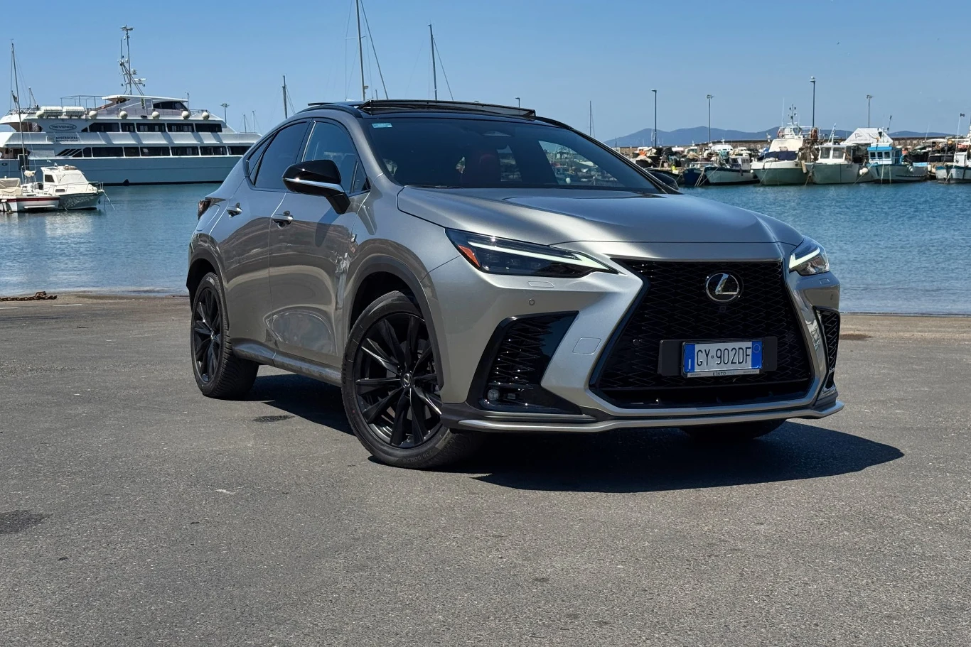 Srebrny samochód Lexus NX zaparkowany na tle przystani z jachtami i łodziami zacumowanymi przy nabrzeżu, w słoneczny dzień.