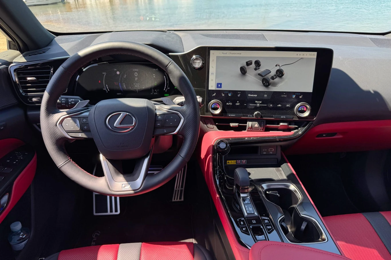 Kokpit nowoczesnego samochodu marki Lexus z dużym ekranem dotykowym na konsoli centralnej, elegancką kierownicą z emblematem marki, sportowymi pedałami oraz czerwono-czarną tapicerką.