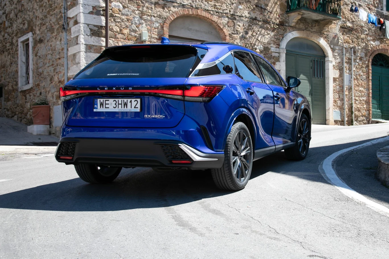 Niebieski SUV marki Lexus zaparkowany na wąskiej, asfaltowej ulicy kamiennego miasteczka, w tle widoczne stare mury budynków i zamknięte zielone drewniane drzwi.