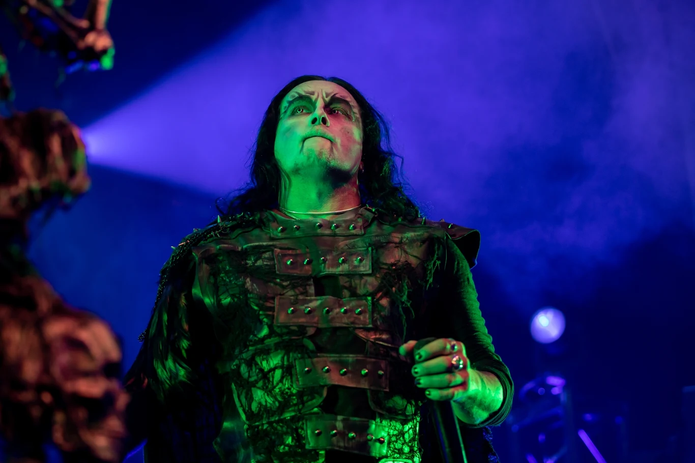 Dani Filth (Cradle of Filth) w akcji Mężczyzna w mrocznym scenicznym makijażu i kostiumie z metalowymi elementami, oświetlony zielonym światłem podczas koncertu, z wyrazistym, poważnym wyrazem twarzy, w tle widoczne kolorowe światła sceniczne.