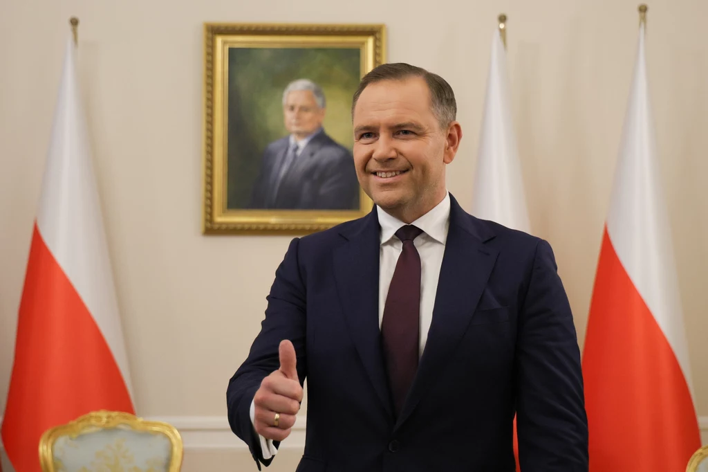 Prezydent elekt Karol Nawrocki Mężczyzna w garniturze uśmiecha się i pokazuje uniesiony kciuk, w tle widoczne są dwie polskie flagi oraz portret mężczyzny na ścianie.