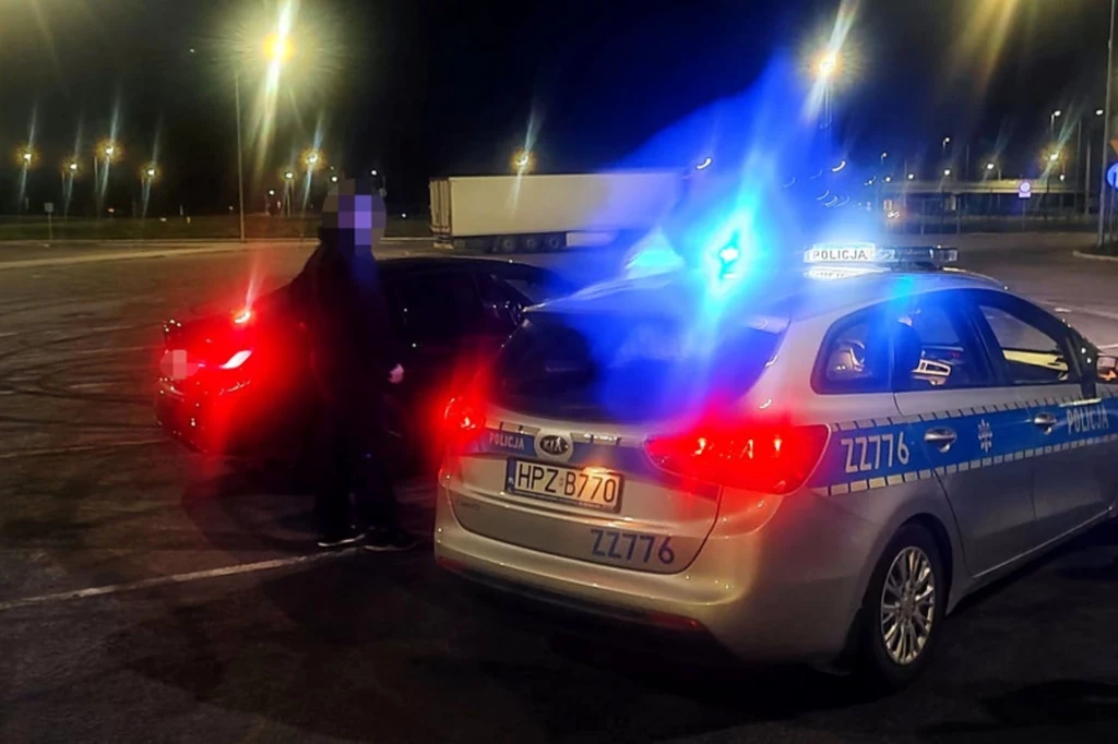 Policjanci ukarali młodego mężczyznę za drift. Policyjny radiowóz z włączonymi sygnałami świetlnymi stoi obok czarnego samochodu osobowego na pustym, nocnym parkingu, obok pojazdów stoi policjant oraz druga postać, widoczne są światełka latarni oraz ciężarówka w tle.