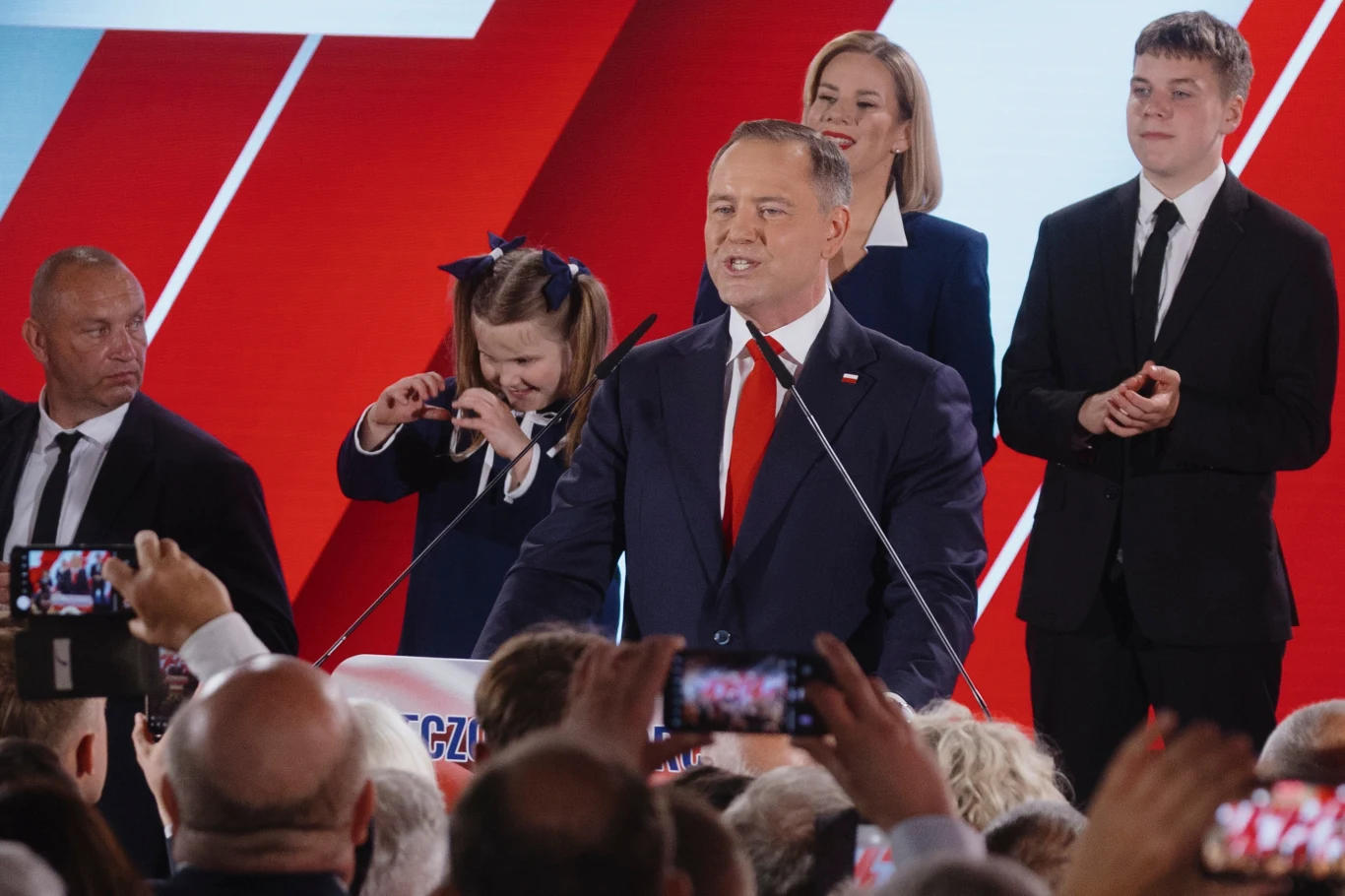 Karol Nawrocki podczas wieczoru wyborczego po drugiej turze wyborów prezydenckich 2025 Polityk w garniturze przemawia do tłumu podczas wiecu wyborczego, otoczony przez współpracowników, ochroniarza i rodzinę, w tle widoczne są biało-czerwone elementy dekoracyjne.