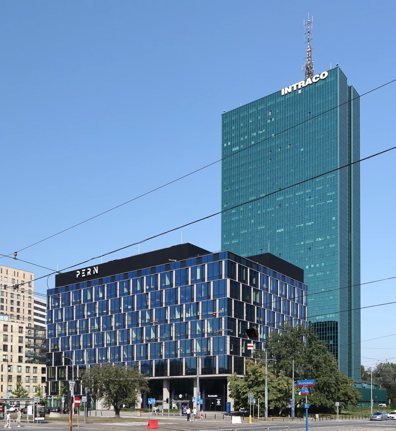 Intraco to dla Warszawiaków to punkt orientacyjny. Dla najemców – luksus w środku bloku wschodniego. Dwa nowoczesne biurowce na tle niebieskiego nieba, większy wieżowiec pokryty zielonym szkłem z napisem Intraco na szczycie, niższy budynek z napisem PERN, wokół znajdują się drzewa i fragmenty ulicy.