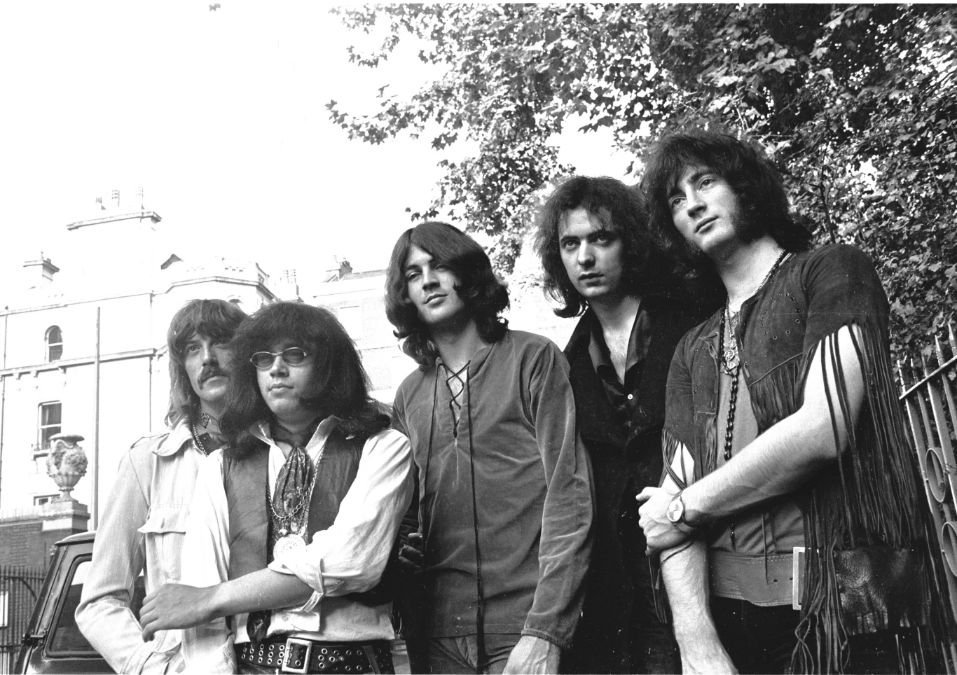 Deep Purple w 1970 r. - od lewej: Jon Lord, Ian Paice, Ian Gillan, Ritchie Blackmore i Roger Glover Pięciu młodych mężczyzn stoi razem na tle budynku i drzew, ubrani w styl typowy dla lat 70., niektórzy mają długie włosy i okulary przeciwsłoneczne, atmosfera jest swobodna i naturalna.