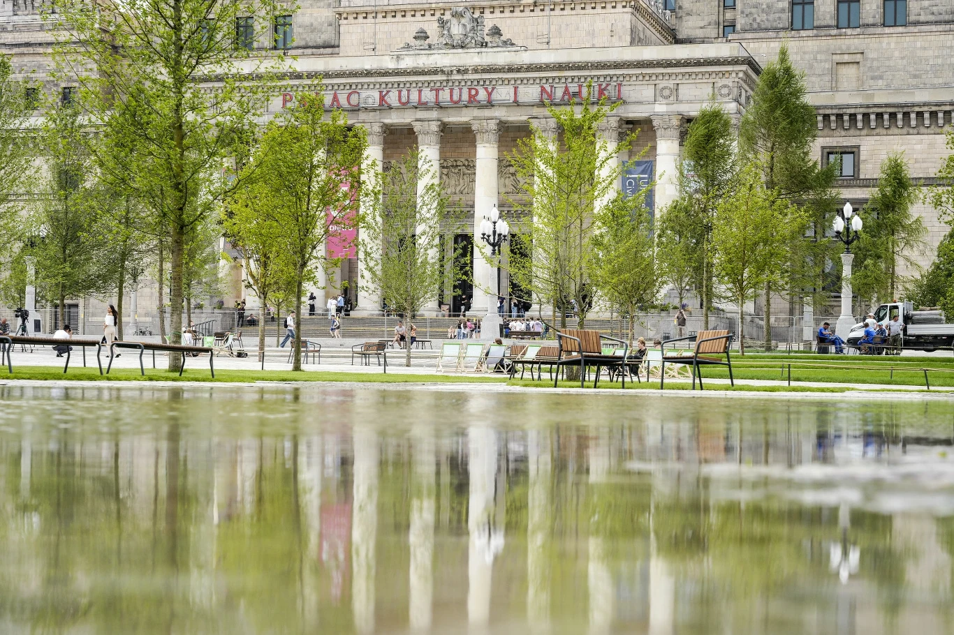 Pałac Kultury i Nauki w Warszawie widoczny zza fontanny i młodych drzew, na pierwszym planie odbicia w wodzie, w tle ławki, zieleń i majestatyczna fasada budynku.
