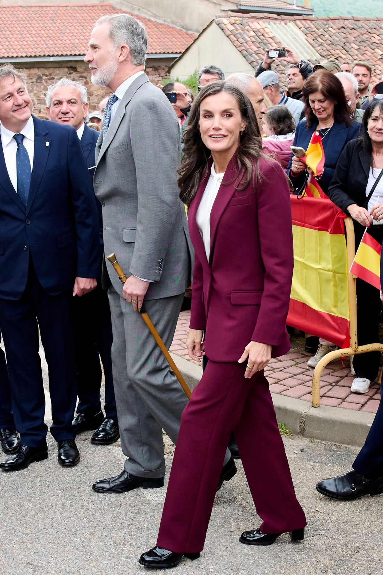 Królowa postawiła na cięższe obuwie w połączeniu z oversizowym garniturem Para uśmiechniętych osób ubranych elegancko idzie razem po ulicy, w tle kilku mężczyzn w garniturach oraz tłum ludzi z flagą Hiszpanii, atmosfera oficjalna i podniosła.