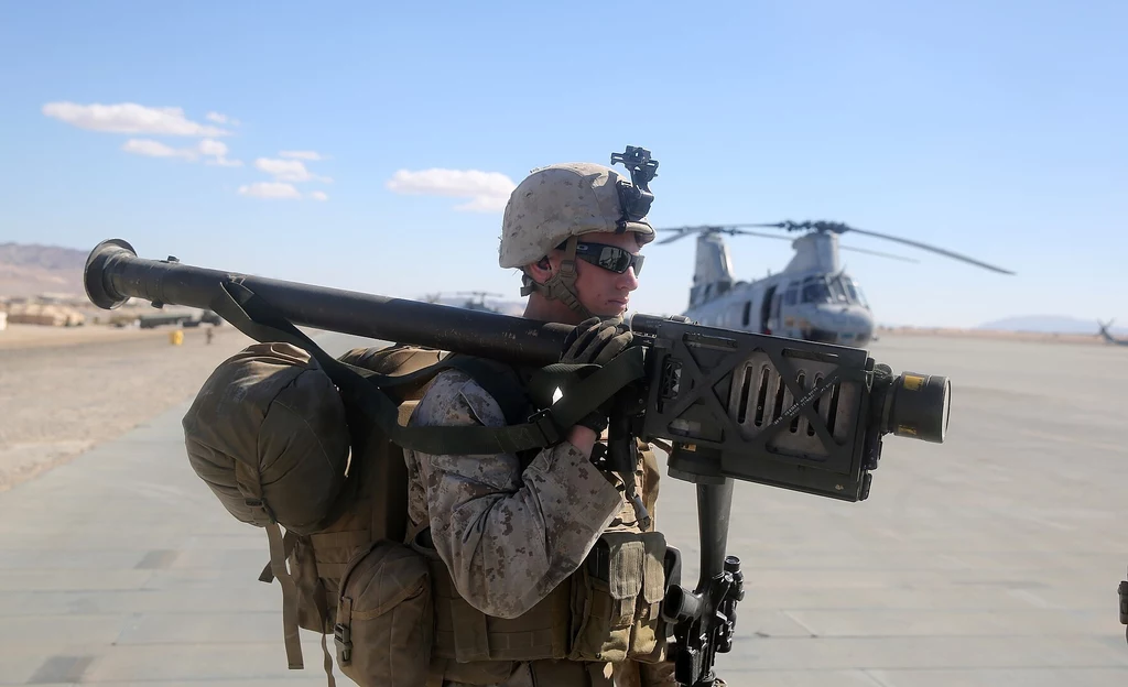 Wyrzutnia FIM-92 Stinger Żołnierz w mundurze polowym niesie na ramieniu przenośny system rakietowy, w tle widoczny śmigłowiec wojskowy stojący na płycie lotniska, jasne niebo i pustynny krajobraz.