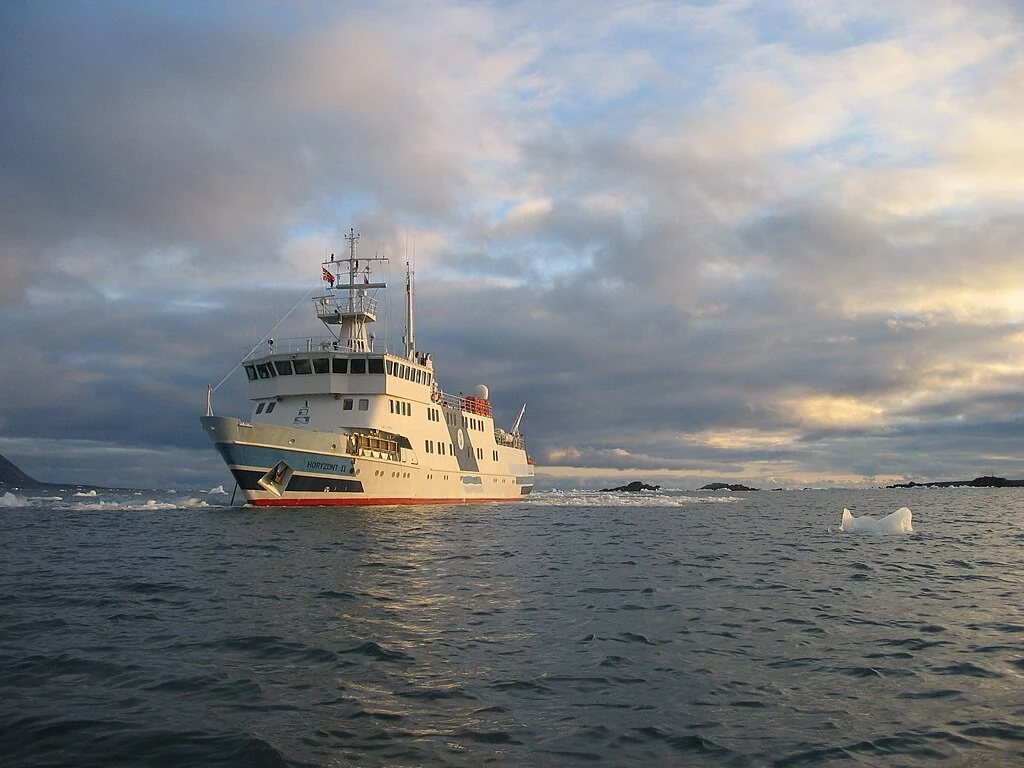 Horyzont II w Zatoce Białego Niedźwiedzia niedaleko Polskiej Stacji Polarnej na Spitsbergenie Duży statek wycieczkowy płynący po spokojnej, zimnej wodzie, na tle chmur i ośnieżonych gór, z fragmentami dryfujących kry lodowej w pobliżu.