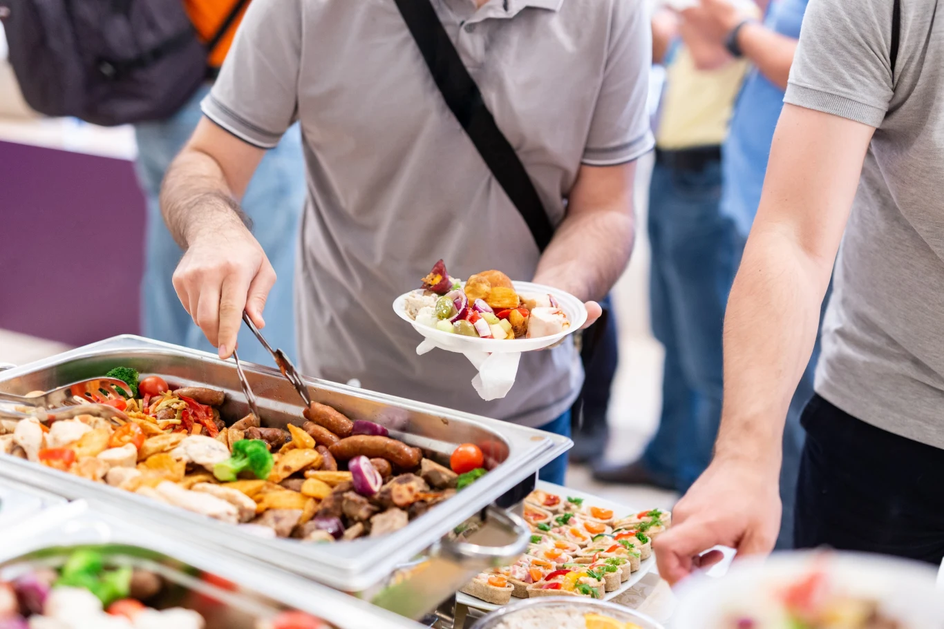 Ręce osób sięgające po jedzenie z bufetu składane na talerze, na stole znajdują się różnorodne potrawy, m.in. mięsa, warzywa i sałatki, atmosfera wskazuje na wydarzenie zbiorowe, np. lunch firmowy lub przyjęcie.