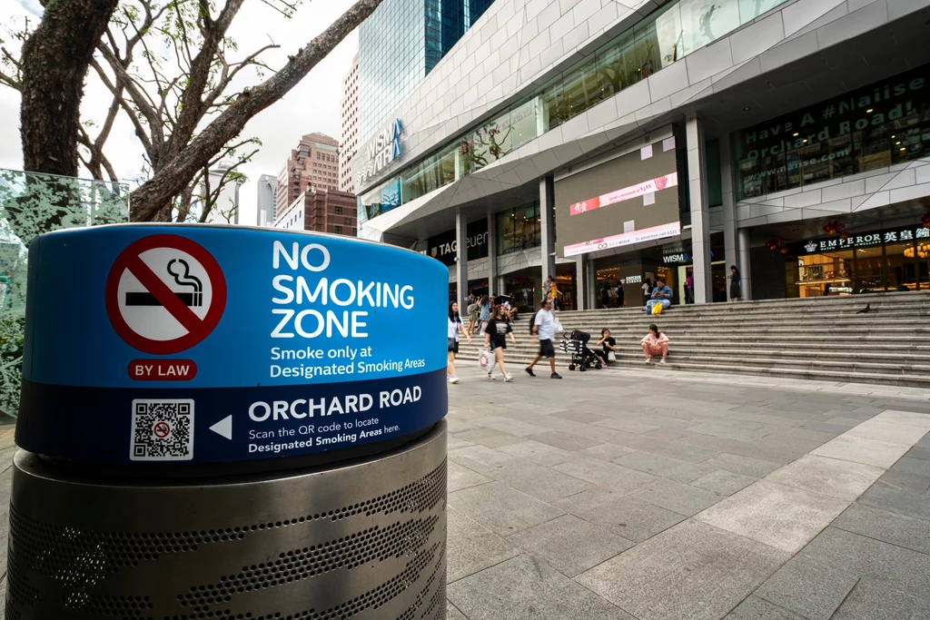 Singapur bardzo poważnie podchodzi do zakazów. Za palenie w nieodpowiednim miejscu grozi nawet 6 tys. zł kary Duży znak z zakazem palenia na centralnym placu handlowym przy Orchard Road, nowoczesne budynki w tle, przechodnie idący w różnych kierunkach, miejsce czyste i uporządkowane