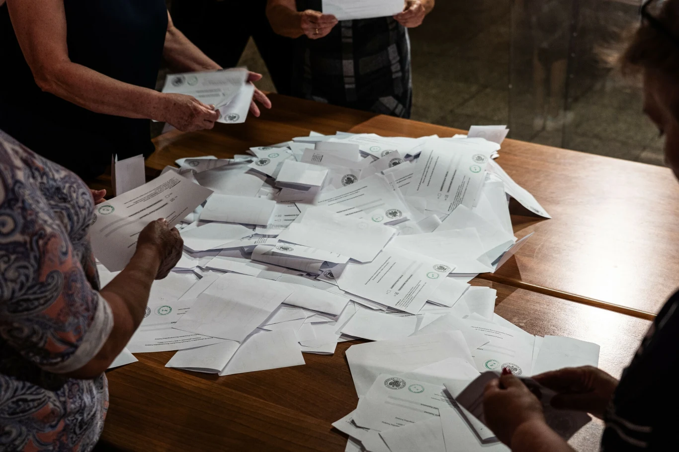 Jeden z protestów wyborczych dotyczy czerwonych korali Ręce kilku osób sortujących lub liczących dużą liczbę kopert, prawdopodobnie związanych z procesem wyborczym, rozrzuconych na drewnianym stole.