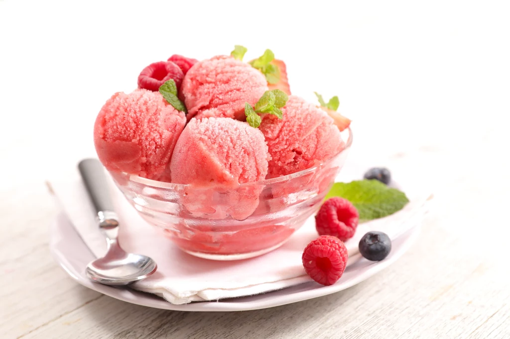 Lody z truskawek, banana i jogurtu to świetny pomysł na szybki i zdrowy deser dla wnuków Porcja malinowego sorbetu podana w szklanej miseczce, ozdobiona świeżymi malinami, jagodami, listkiem mięty i serwowana na białym talerzyku z łyżeczką.