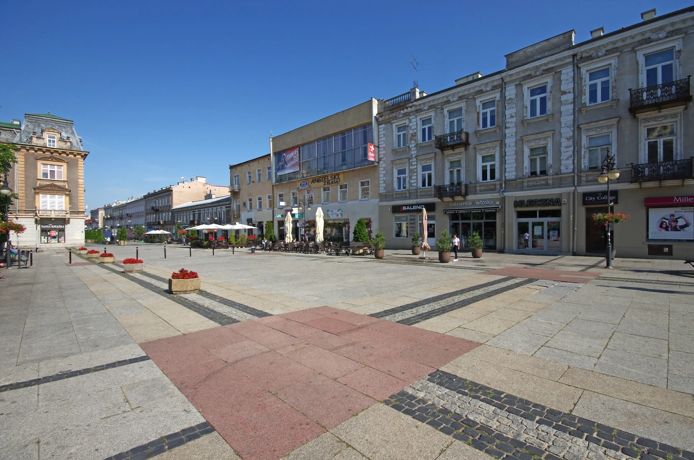 Pusta przestrzeń miejska otoczona zabytkowymi kamienicami, ustawione na zewnątrz kawiarniane stoliki, kilka donic z kwiatami oraz sklepowe witryny po prawej stronie.