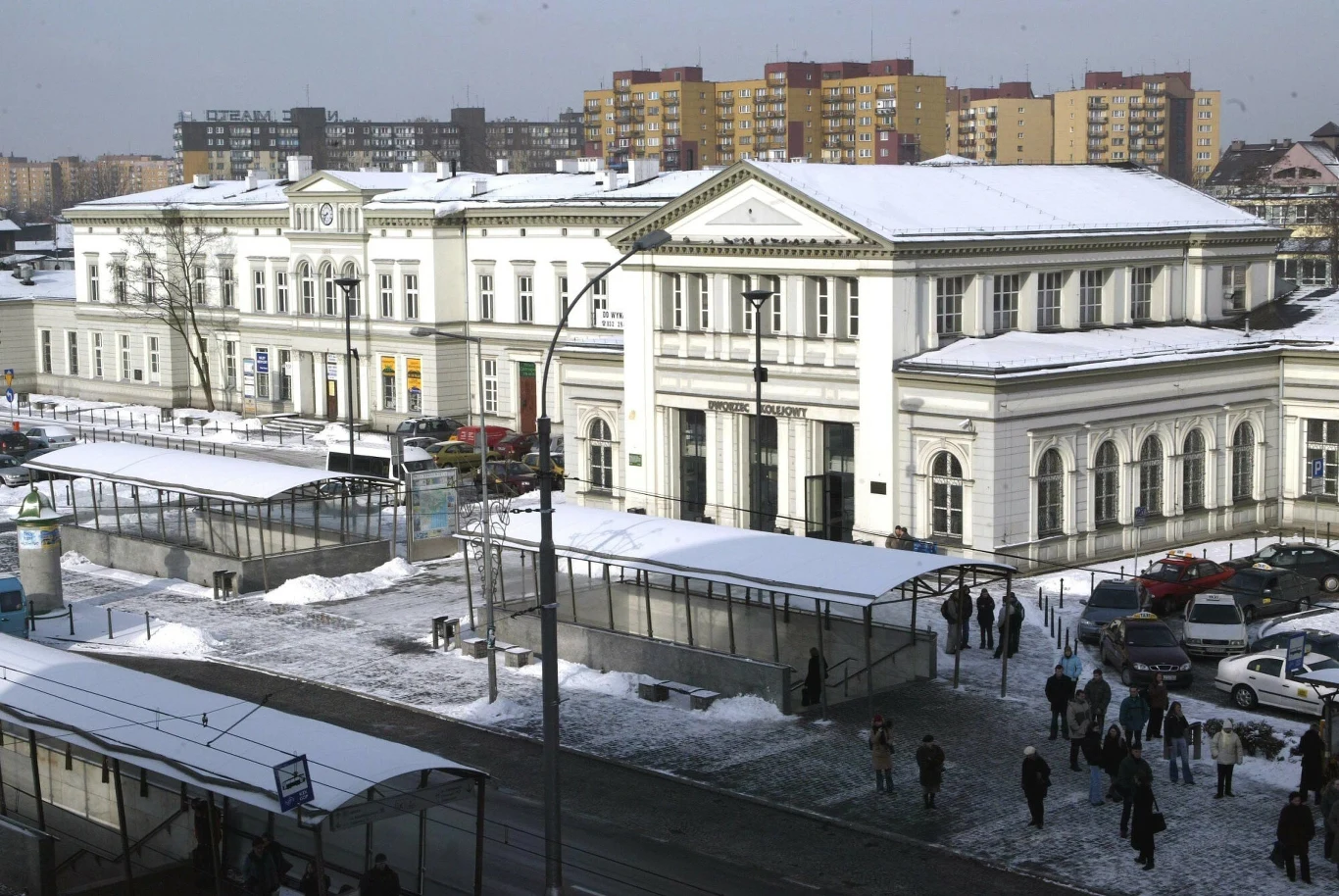 Historyczny budynek dworca kolejowego na tle zimowego miasta, z przykrytymi śniegiem dachami, otoczony przez przechodniów, samochody oraz bloki mieszkalne w tle.