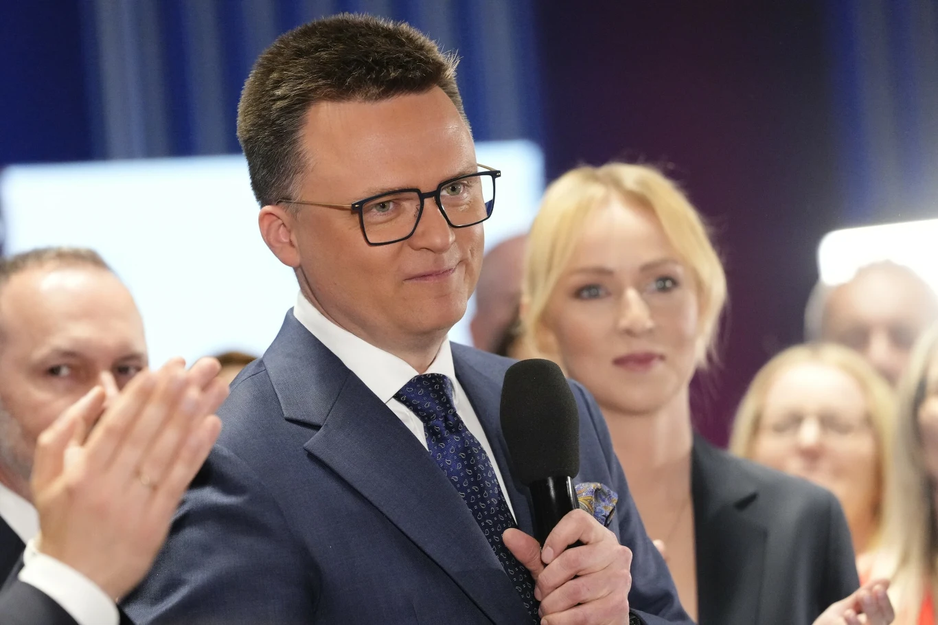 Mężczyzna w garniturze i okularach trzyma mikrofon, wokół niego grupa osób, wszyscy skupieni i elegancko ubrani, scena w jasnym oświetleniu.
