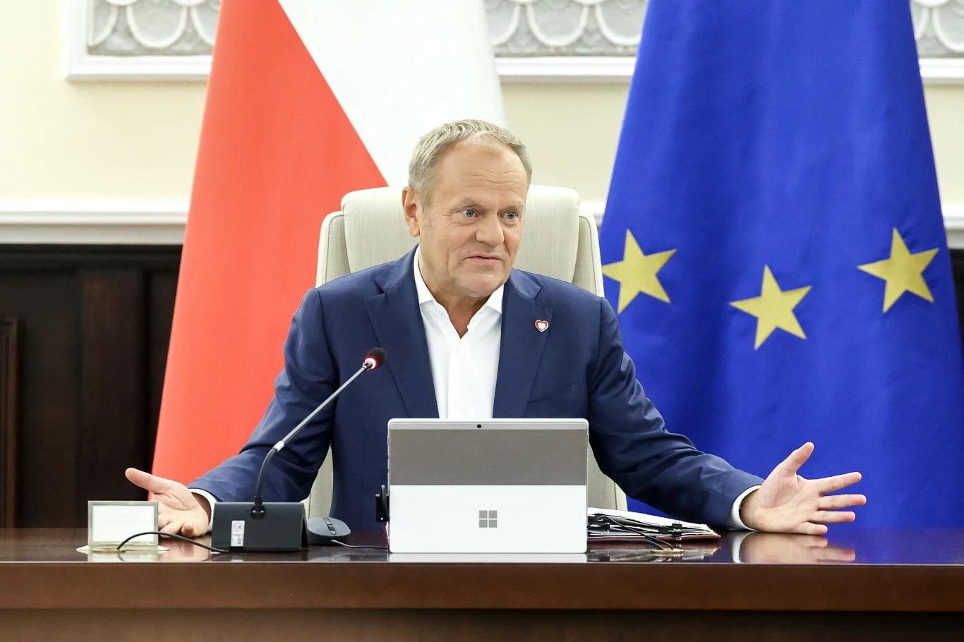 Mężczyzna w garniturze siedzi przy dużym, drewnianym stole, przed nim urządzenie elektroniczne i mikrofon, w tle widać flagę Polski oraz flagę Unii Europejskiej.