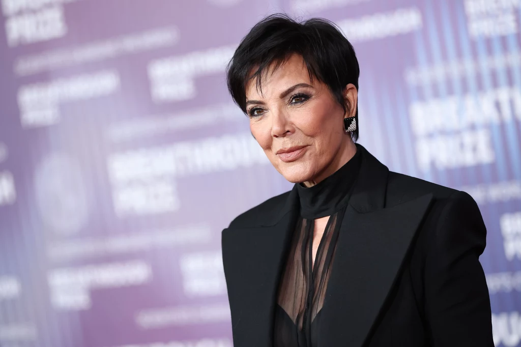 Jenner wstaje o 4:30, a dzień zaczyna od czytania najnowszych wiadomości ze świata Kobieta w krótkich, ciemnych włosach, w eleganckim czarnym garniturze z przezroczystą koszulą, stoi na tle fioletowej ścianki z rozmytymi napisami, patrząc lekko w bok z delikatnym uśmiechem.