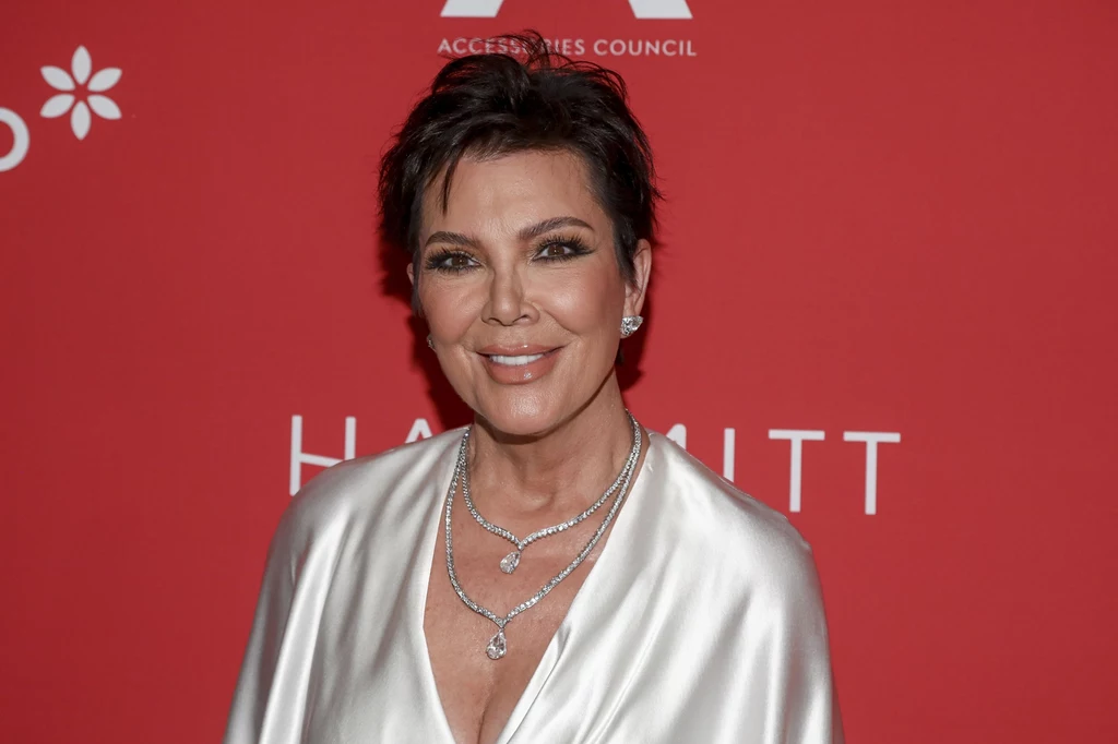 Matka klanu Jenner-Kardashian stawia na planowanie. To pozwala jej zachować porządek podczas wykonywania obowiązków Kobieta w eleganckiej, perłowej sukni, z krótkimi, ciemnymi włosami, uśmiechnięta, stoi na tle czerwonej ścianki z białymi napisami oraz logotypami, nosi wyrazistą biżuterię i makijaż podkreślający rysy twarzy.
