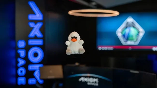 "Joy", czyli wskaźnik zerowej grawitacji dla misji Axiom Mission 4. Pluszowa zabawka w kształcie kaczki unosi się w powietrzu na tle nowoczesnego biura ze znakiem Axiom Space oraz dużym ekranem z kolorową grafiką.
