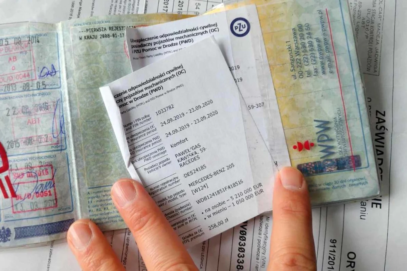 Nowelizacja przepisów dotyczących polis OC ułatwi kierowcom wypowiadanie umów Dowód rejestracyjny pojazdu wraz z dokumentami ubezpieczeniowymi oraz zaświadczeniem, trzymany w ręce na tle innych papierów.