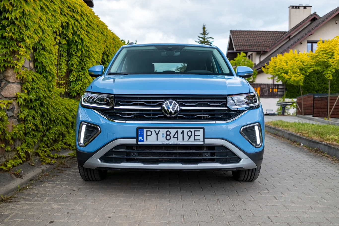 Nowoczesny niebieski samochód marki Volkswagen zaparkowany na brukowanej ulicy między zielonymi żywopłotami a domami jednorodzinnymi, widok od frontu, tablica rejestracyjna Polski.