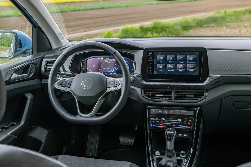 W kabinie odświeżonego Volkswagena T-Cross znajdziemy już nieco więcej nowości Wnętrze samochodu Volkswagen z kierownicą, cyfrowym kokpitem, dużym ekranem dotykowym systemu multimedialnego oraz konsolą centralną z automatyczną dźwignią zmiany biegów.