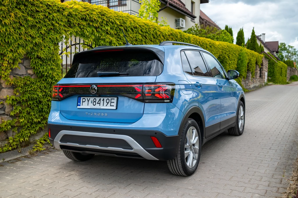 Z tyłu Volkswagen T-Cross po liftingu zmienił się w niewielkim stopniu Błękitny samochód marki Volkswagen zaparkowany tyłem na brukowanej, osiedlowej ulicy, otoczonej zielonym żywopłotem i domami jednorodzinnymi.
