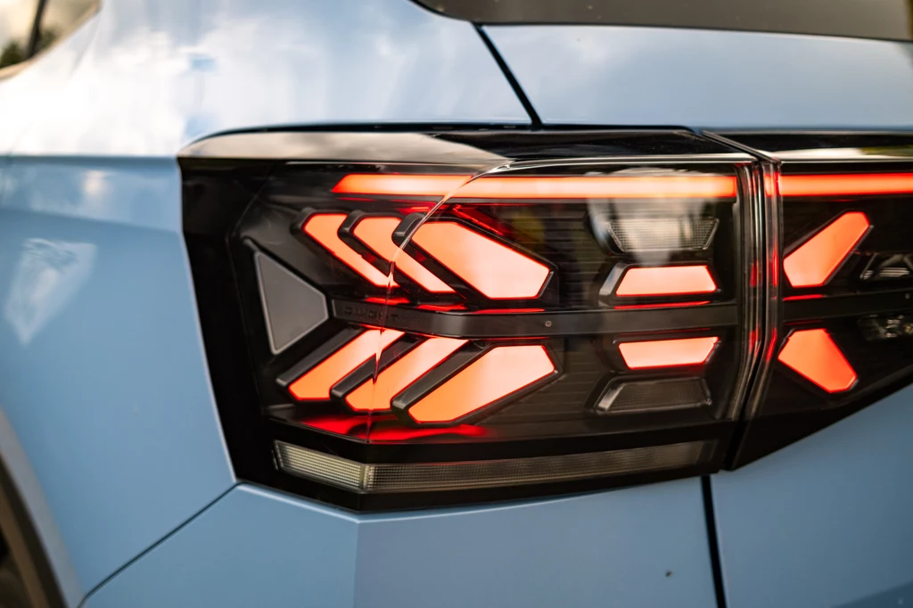 Volkswagen T-Cross Nowoczesna tylna lampa samochodu o charakterystycznym, geometrycznym kształcie i czerwonych światłach LED na tle jasnoniebieskiego lakieru.