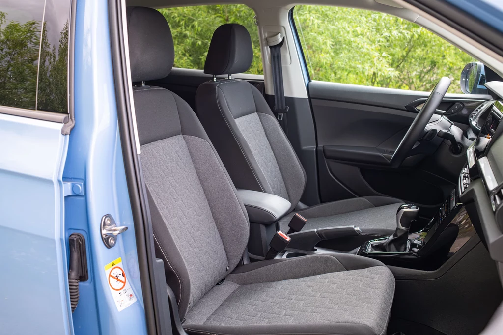 Volkswagen T-Cross Nowoczesne wnętrze samochodu z widocznymi przednimi siedzeniami, deską rozdzielczą oraz elementami drzwi, częściowo otwarte drzwi po stronie kierowcy, jasna tapicerka i detale w czarnym kolorze, widoczna zieleń za szybami samochodu.