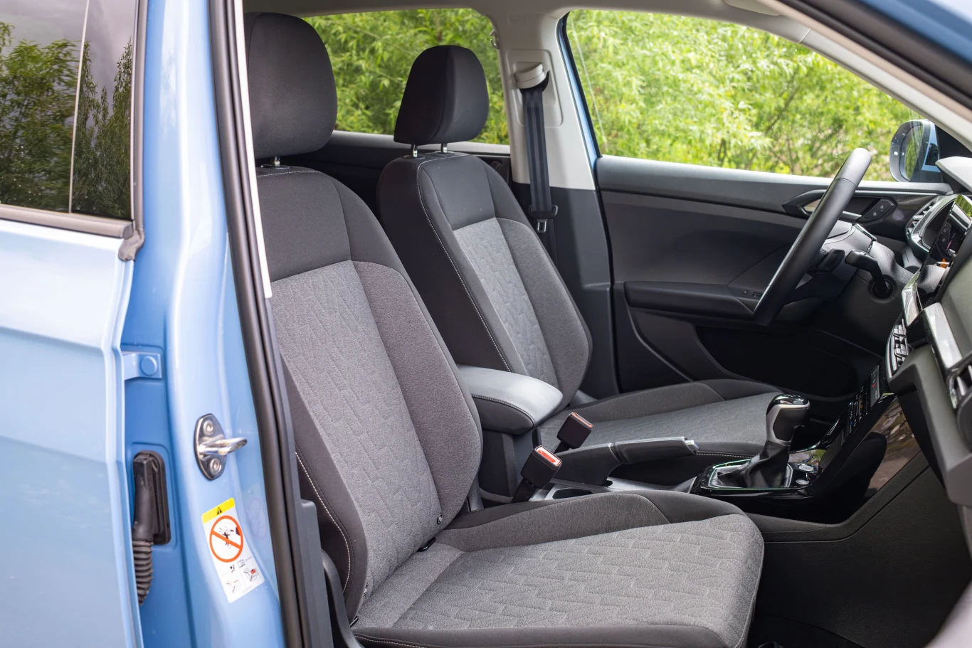 Nowoczesne wnętrze samochodu z widocznymi przednimi siedzeniami, deską rozdzielczą oraz elementami drzwi, częściowo otwarte drzwi po stronie kierowcy, jasna tapicerka i detale w czarnym kolorze, widoczna zieleń za szybami samochodu.