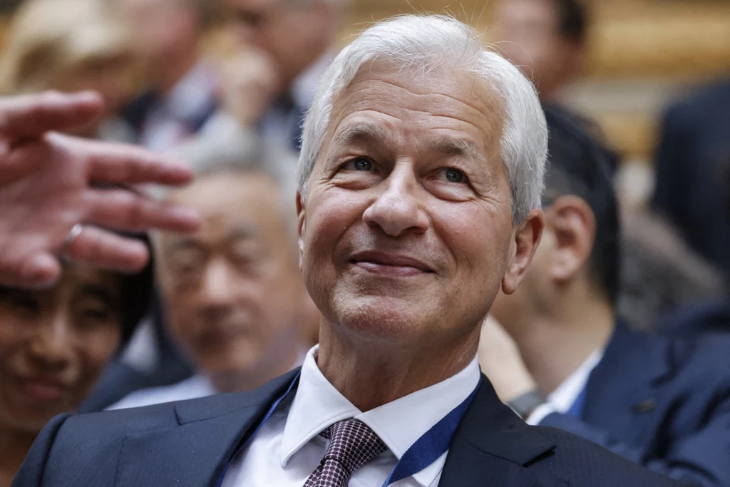 Jamie Dimon Starszy mężczyzna z siwymi włosami w garniturze i krawacie, uśmiechnięty, siedzący wśród innych osób podczas wydarzenia o charakterze biznesowym lub konferencyjnym, w tle nieostre twarze i sylwetki ludzi.