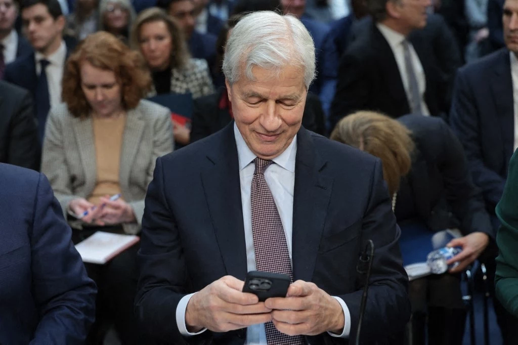 Jamie Dimon w Kongresie Starszy mężczyzna w garniturze z siwymi włosami patrzy na telefon komórkowy, siedząc wśród innych ludzi podczas wydarzenia o formalnym charakterze, w tle widać skupione osoby z notatnikami.