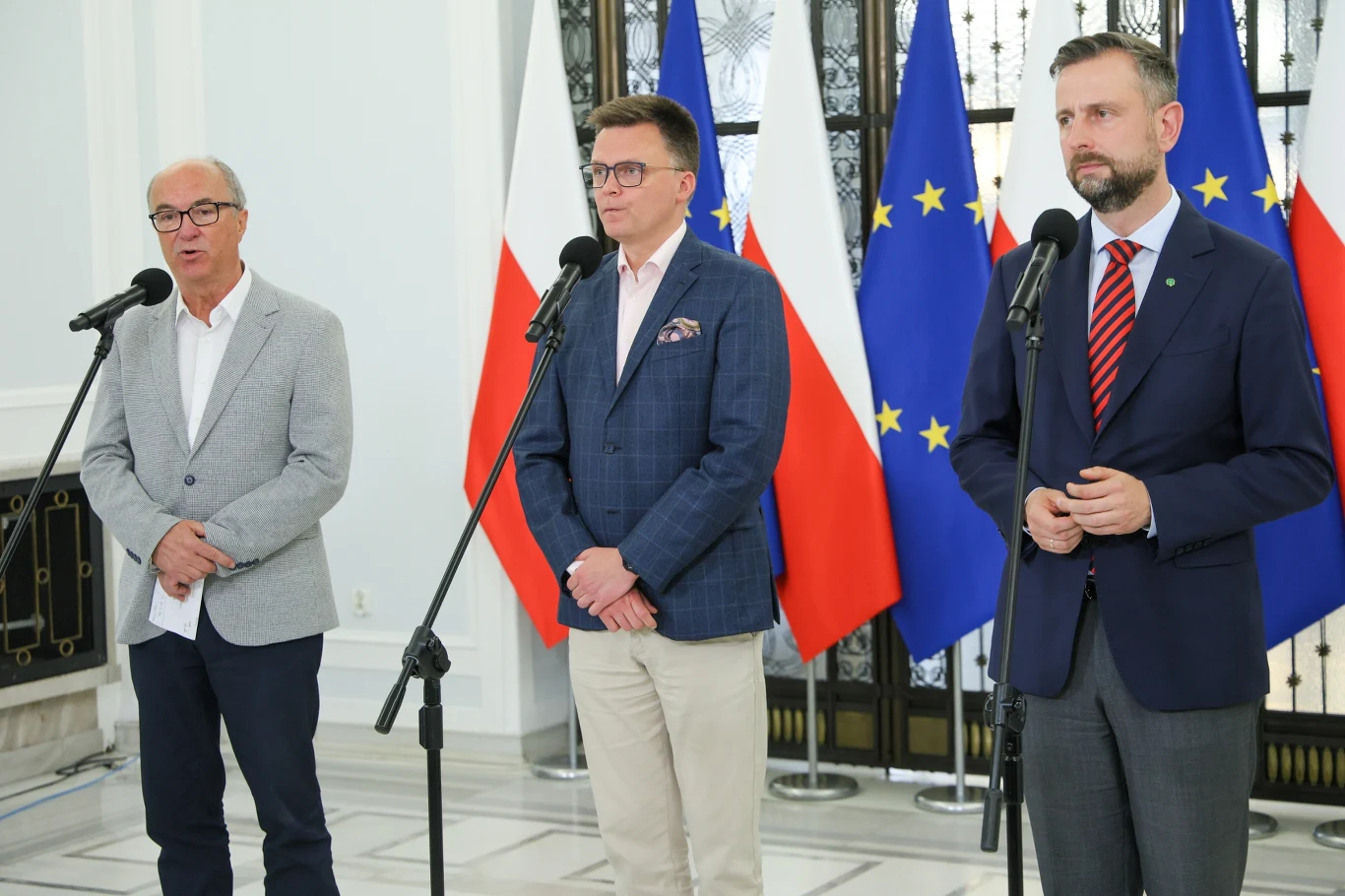 Trzech mężczyzn w garniturach stoi przy mikrofonach w eleganckim wnętrzu, w tle widoczne są flagi Polski oraz Unii Europejskiej.