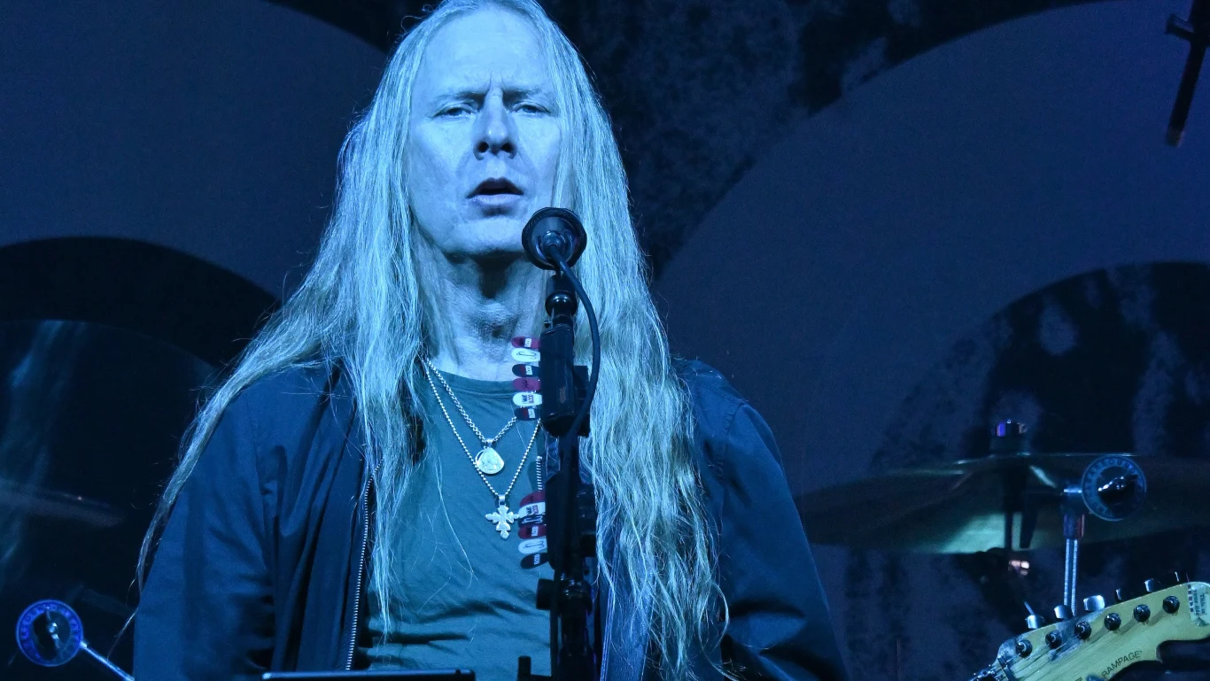 Jerry Cantrell Długowłosy mężczyzna śpiewający do mikrofonu na scenie koncertowej, w tle widoczne bębny i fragment gitary, całość oświetlona niebieskim światłem.