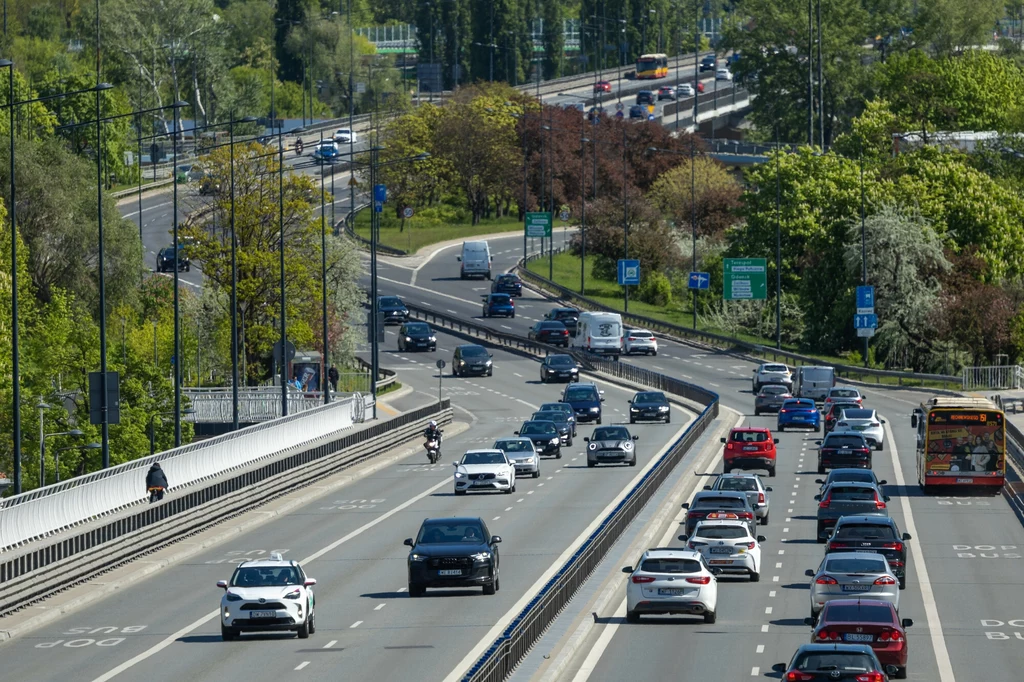 Św. Krzysztof uważany jest za patrona kierowców Duży miejski most z czteropasmową drogą, liczne samochody jadące w obu kierunkach, autobus miejski, zielone drzewa i tablice drogowe w tle, wiosenna pogoda.