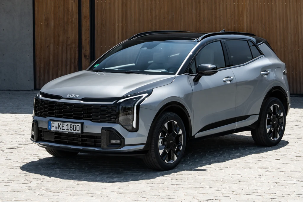 Nowa Kia Sportage Srebrny SUV marki Kia stoi zaparkowany na brukowanym placu przed drewnianą i betonową ścianą, auto posiada charakterystyczne czarne wykończenia oraz nowoczesny design świateł.