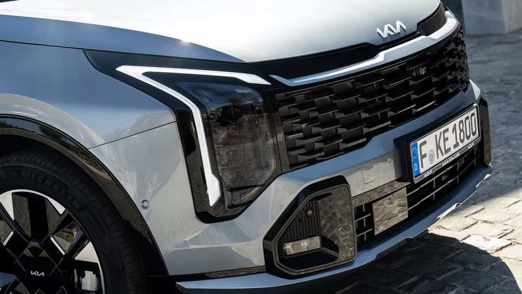 Sporo zmian pojawiło się w przedniej części nowej Kii Sportage. Teraz wygląda znacznie bardziej muskularnie szczególnie w wersji GT-Line z większymi obręczami. Przód nowoczesnego srebrnego samochodu marki Kia z charakterystycznym grillem, nowoczesnym wzorem świateł i widoczną niemiecką tablicą rejestracyjną, stojący na brukowanym podłożu.
