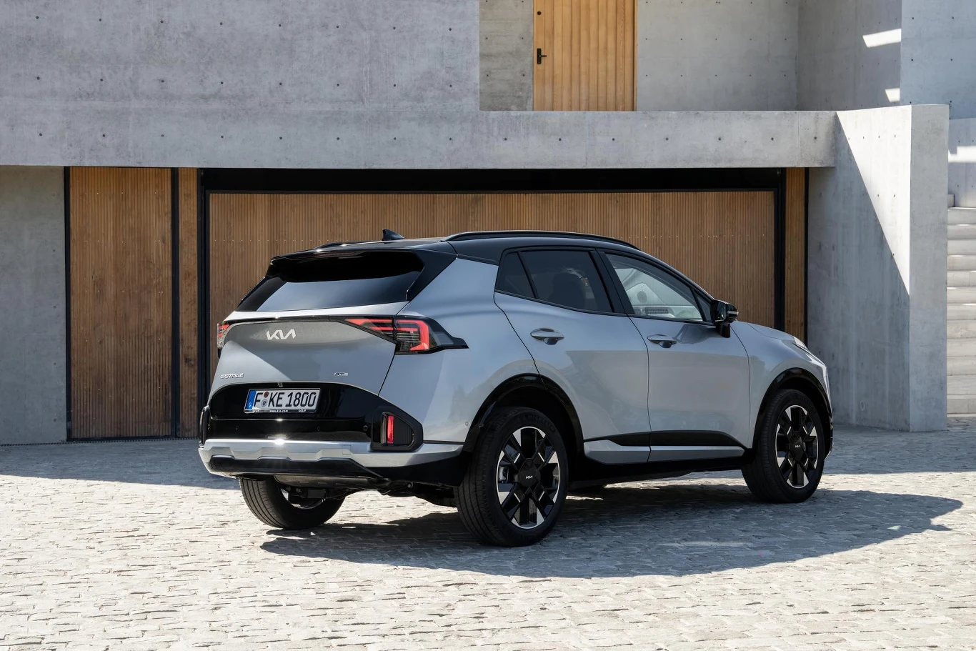 Srebrny samochód Kia Sportage zaparkowany na brukowanym podjeździe przed nowoczesnym budynkiem o betonie i dużych, drewnianych drzwiach, widok z tyłu pod kątem.