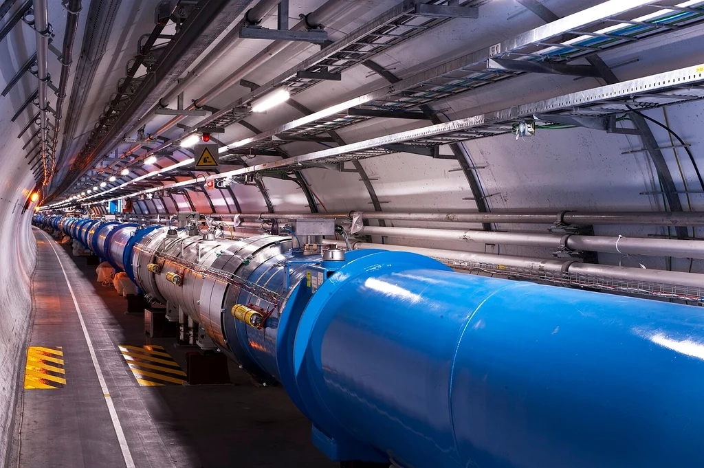 Fragment tunelu Wielkiego Zderzacza Hadronów Długi tunel z metalowymi, cylindrycznymi konstrukcjami zdominowanymi przez niebieską obudowę, liczne przewody oraz oznaczenia ostrzegające o zagrożeniu elektrycznym, całość oświetlona lampami biegnącymi wzdłuż sufitu.