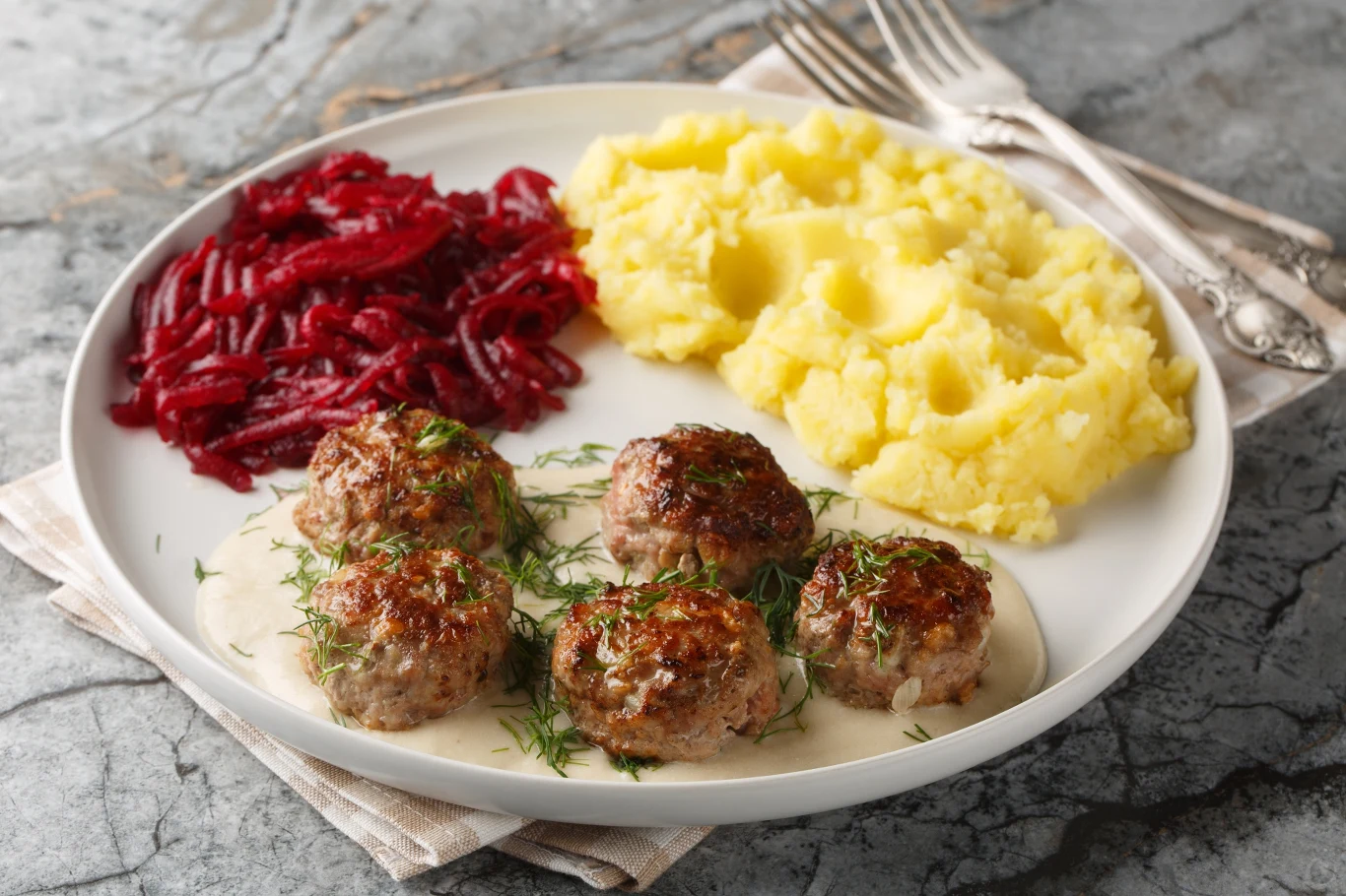 Porcja pieczonych klopsików w sosie koperkowym udekorowanych świeżym koperkiem, podana z puree ziemniaczanym oraz tartymi buraczkami na białym ceramicznym talerzu, sztućce w tle.