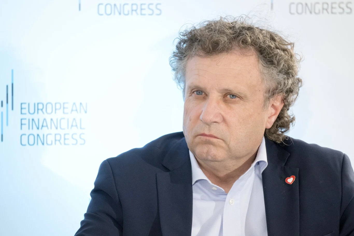 Jacek Karnowski ogłosił swój powrót do Platformy Obywatelskiej Mężczyzna w średnim wieku w garniturze siedzi na tle panelu z napisem 'European Financial Congress', wyraz twarzy poważny, przypinka w kształcie serca na klapie marynarki.