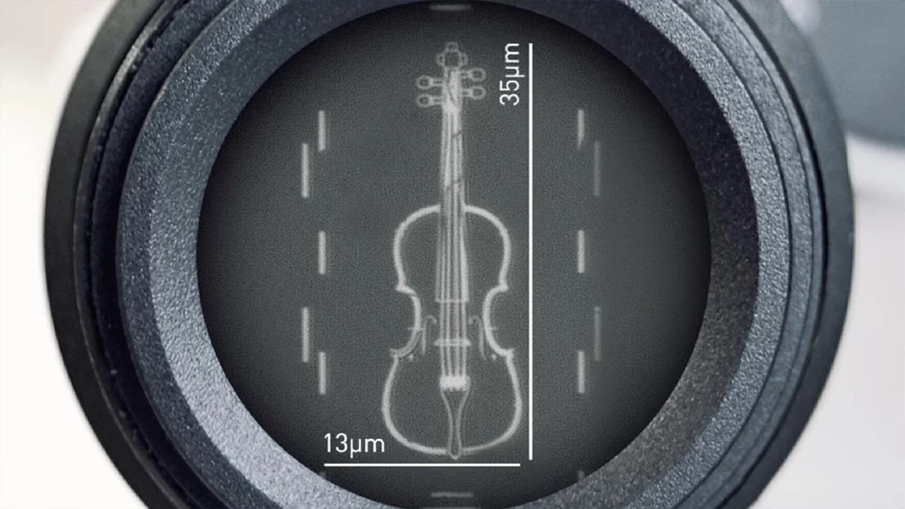 Wnętrze okrągłego obiektywu mikroskopu, w którym widoczny jest miniaturowy rysunek skrzypiec o wymiarach 35 mikrometrów na 13 mikrometrów, z naniesionymi wymiarami i podziałkami pomocniczymi.