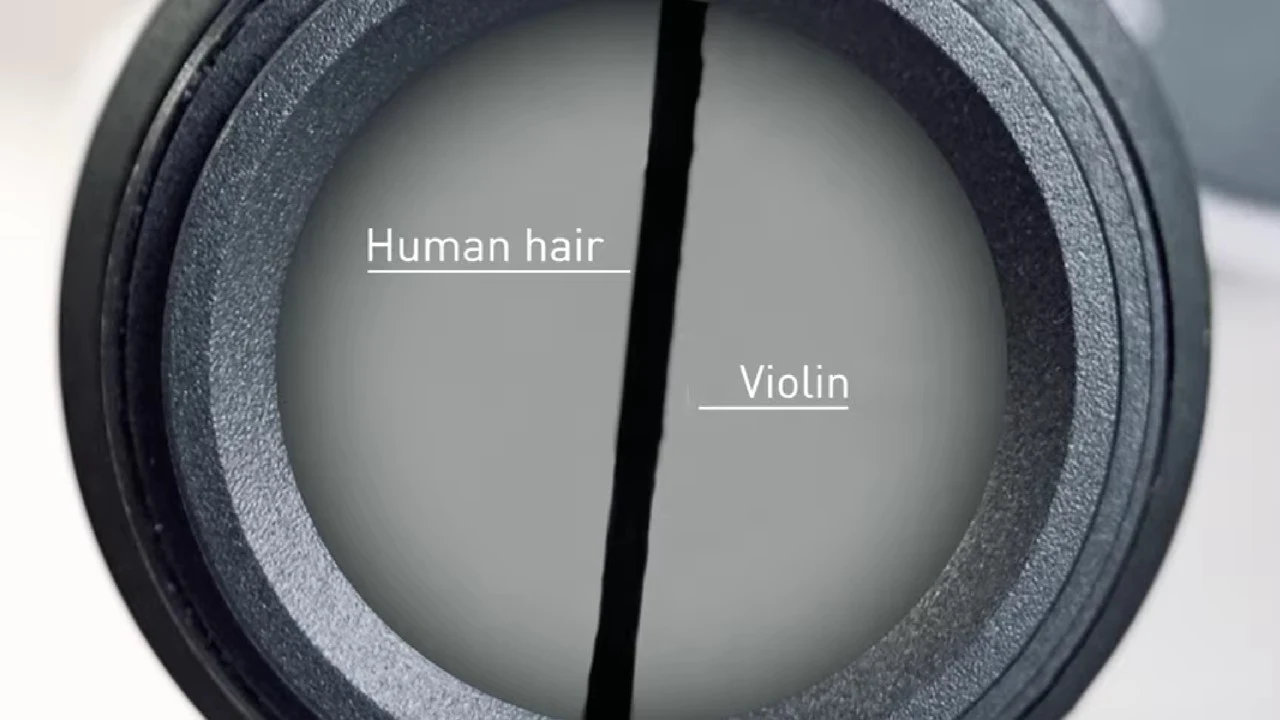 Porównanie grubości ludzkiego włosa i włókna z wiolinowego smyczka pod mikroskopem, oba oznaczone opisami, w okrągłym polu widzenia soczewki.